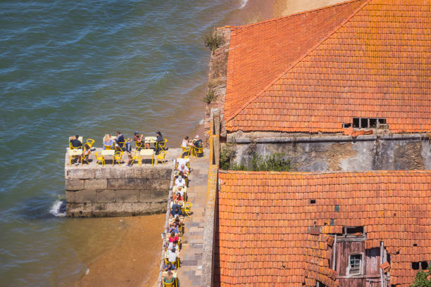 Scenic view of the picturesque seaside village Azenhas do Mar in Portugal, with traditional white houses and red roofs perched on dramatic cliffs above the Atlantic Ocean. The natural seawater pool and beach area are visible below, with people enjoying a sunny day. Captured on a clear summer afternoon.