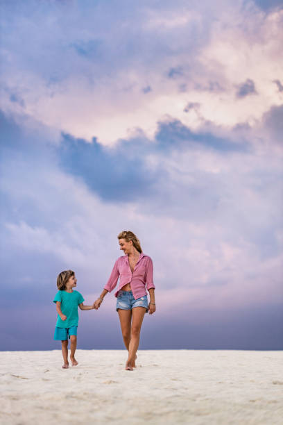 Happy mother and son holding hands and communicating while taking a walk on the beach under moody sky. Copy space.