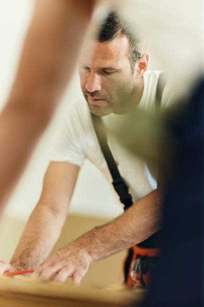 A determined self-builder in a hard hat and work gloves drives screws into an OSB panel with a cordless drill, securing it to the wooden frame of his home
