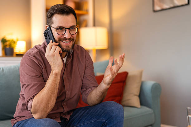 istock Young man is sitting on a sofa at home, engaged in a phone conversation. The scene captures a moment of communication, relaxation, or multitasking in a comfortable living space. 2202036789
