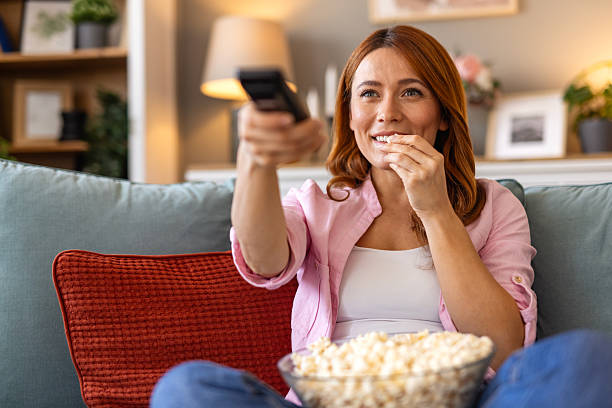 istock A woman is sitting on a cozy sofa, enjoying a TV show while eating popcorn. The image captures a perfect moment of leisure, ideal for themes related to relaxation, entertainment, and home life. 2202037471