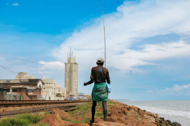 Colombo, Sri Lanka - September 12, 2024: A local fisherman stands on the rocky shore of Colombo, casting his fishing line into the sea under a clear blue sky.