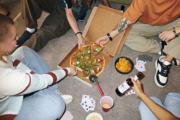 Group of people sitting on floor enjoying pizza from box and beverages while playing cards. Relaxed social setting with snacks and casual atmosphere
