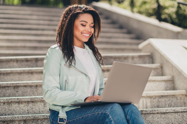 istock Young woman enjoying daylight on urban steps with a smile, wearing a stylish jacket while working on her laptop outdoors during a warm day. 2202045034