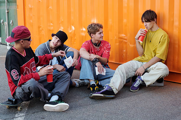 Group of four teenagers sitting outside near storage containers while enjoying drinks and snacks, casually interacting with one another and sitting on skateboards