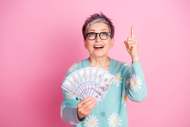 istock Happy elderly woman holding dollar bills with a surprised expression on a pink background 2202045603