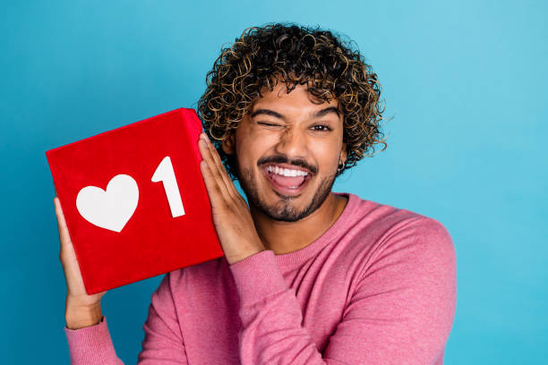 istock Smiling and playful young man in a pink pullover holding a red block with a heart sign against a blue background 2202047871