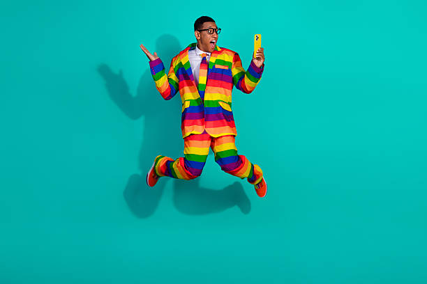 istock Energetic guy in a rainbow suit jumps joyfully on a teal background while holding a vibrant yellow smartphone. 2202055546