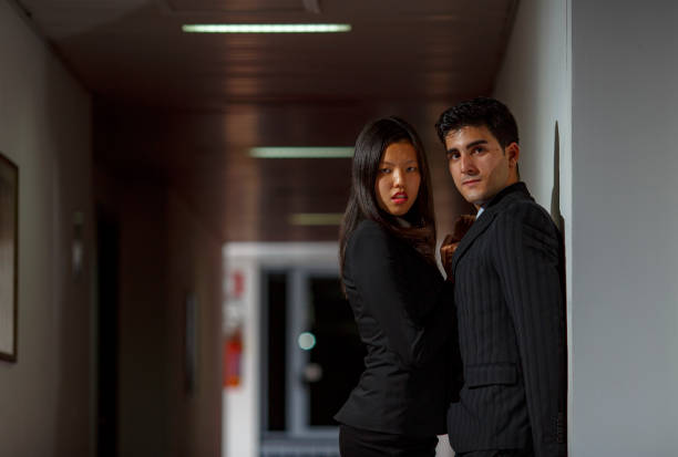 A man and a woman stand confidently in front of an ornate wooden door, dressed in stylish formal attire. The woman wears a sophisticated dress with ruffled accents, while the man sports a sharp suit.