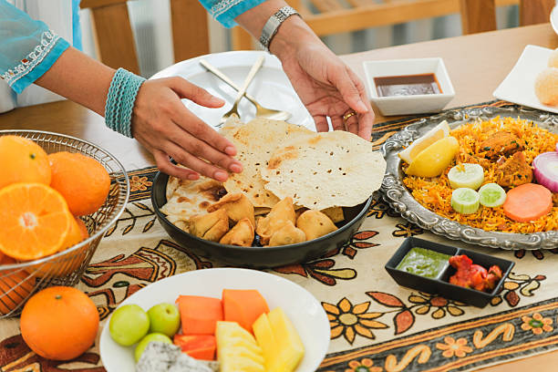 A festive Indian meal featuring samosas, papad, naan, and biryani, served on an ornate tablecloth. A woman's hands arrange the food, accompanied by chutneys, fruits, and fresh garnishes.