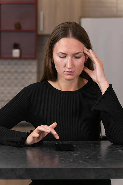 istock Young woman sitting at a kitchen table, using her smartphone while holding her head in frustration, feeling stressed and exhausted from the demands of working from home. Vertical photo 2202061466
