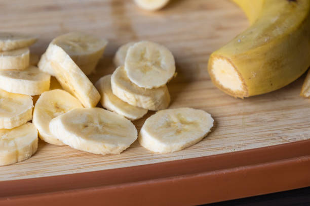 Freshly sliced banana pieces are served in a clear glass bowl. A metal fork rests on the wooden cutting board next to the bowl, ready to enjoy this simple and healthy snack.
