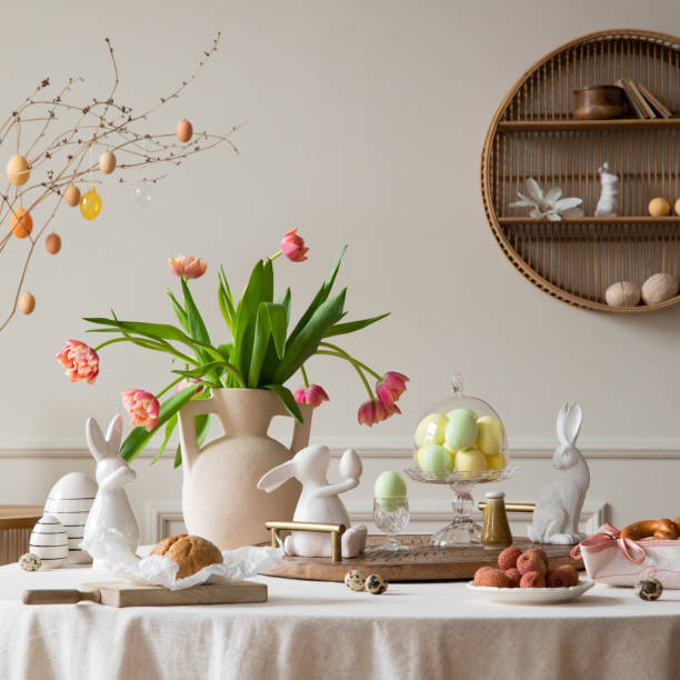 istock Creative composition of spring easter dining room interior with round table, vase with tulips, colorful eggs, wooden trace, bread, easter bunny, sculpture and personal accessories. Home decor Template 2202068950