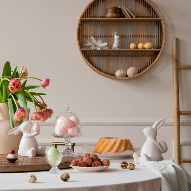 istock Interior design of easter dining room interior with round table, shelf on wall, colorful eggs, easter cake, hare sculptures, vase with tulips, lychee and personal accessories. Home decor. Template. 2202069020