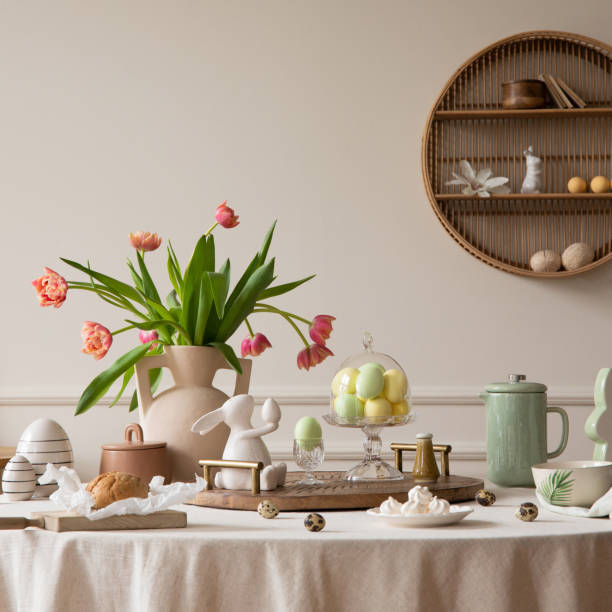 istock Creative composition of easter dining room interior with round table, vase with tulips, colorful eggs, pitcher, wooden trace, bread, hare sculpture and personal accessories. Home decor. Template. 2202069021