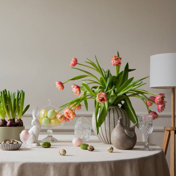 istock Interior design of easter dining room with colorful eggs, white hare sculptures, vase with tulips, plants, lamp, beige wall with stucco, gray hen and personal accessories. Home decor. Template. 2202069097