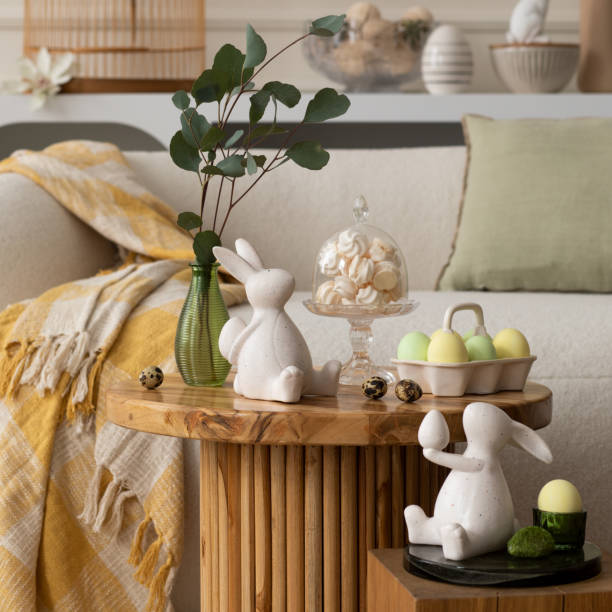 istock Spring composition of living room interior with round coffee table, beige sofa, easter bunny sculpture, colorful easter eggs,  yellow plaid, meringues and personal accessories. Home decor. Template. 2202074676