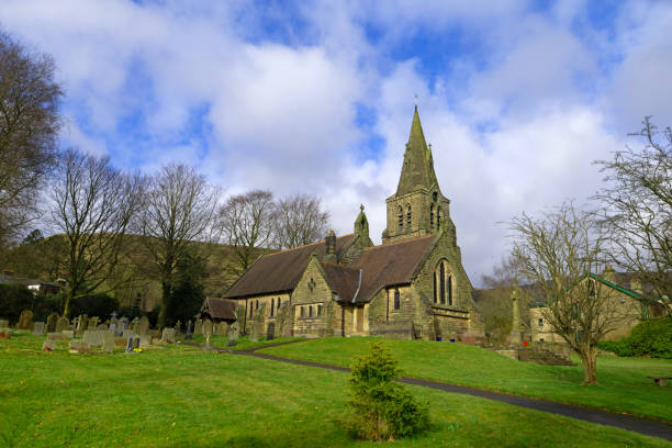 Gravel path through dry stone wall and iron gate welcoming worshippers into picturesque Cotswold country churchyard and idyllic rural church with arch door, traditional stone slate roof and short square tower under blue summer skies. ProPhoto RGB profile for maximum color fidelity and gamut.