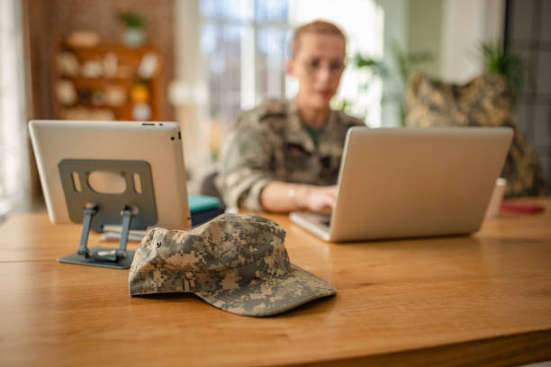 A young woman in military uniform is seated at a home office desk, engaged with a laptop while a tablet rests nearby. A camouflage cap is placed in the foreground, signifying her service.
