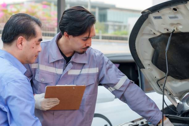 istock Male insurance officer in a gray uniform is pointing at a car engine while explaining accident coverage to a client. Professional man is assessing damage and handling insurance paperwork. 2202108160