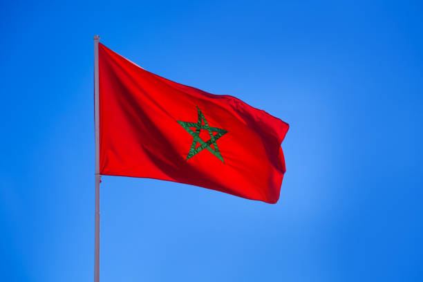 A vibrant Moroccan flag prominently displayed waving against a bright, clear blue sky. Showcasing patriotism and national pride, this image captures the beauty and symbolism of Morocco's national emblem.