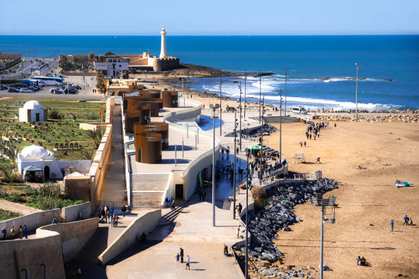 Beautiful coastal scene in Rabat, Morocco, featuring a historic lighthouse, modern structures, and people enjoying the landmark near the ocean.