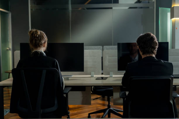 istock Two professionals sitting at modern office desk facing computer screens. Glass partitions and dim lighting creating a focused work atmosphere. Business setting illustrating teamwork and corporate 2202118512