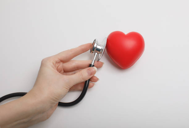 Hand holding stethoscope checking red heart on white background, symbolizing cardiology, healthcare, heart disease prevention, and medical diagnostics. Concept of heart health, love, and wellness.