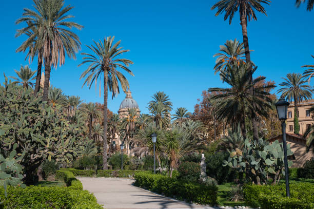 Villa Bonanno public garden in Palermo, Sicily, featuring tall palm trees, green pathways, and exotic plants. A historic dome structure rises in the background under a clear blue sky, creating a serene and picturesque setting.