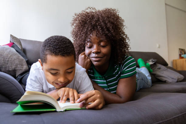 A female babysitter of African descent is indoors with a young Caucasian boy. They are wearing casual clothing. They are reading a storybook together. The boy is on the girl's lap.