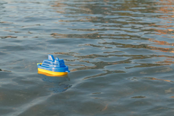 A blue and yellow plastic toy boat floats on a calm water surface. Gentle ripples reflect the sunlight, creating a peaceful and playful scene.