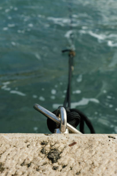 Close-up of a metal mooring ring securing a black rope on a stone dock by the water, with soft waves in the background.