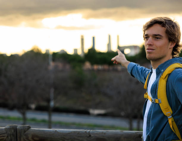 A young adventurer with a yellow backpack enthusiastically points towards Madrid's skyline during sunset. His expression reflects excitement and appreciation for the view.