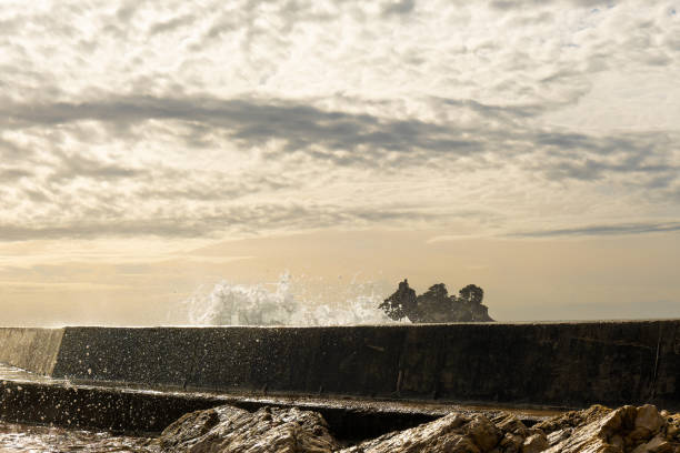 A dramatic wave crashes against a seawall at sunset, with water droplets suspended in the air and a rocky shoreline in the foreground.