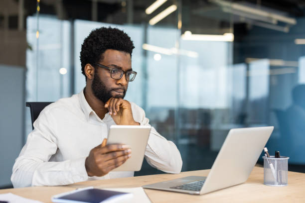 istock Male thinking businessman working with laptop and tablet computer inside office, man in business shirt viewing online financial report, reading from monitor screen 2202135776