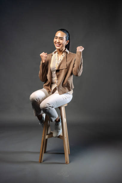 A woman sitting on a stool in a studio environment demonstrates happiness with a cheerful gesture. The warm lighting and neutral background emphasize her expression and bring focus to the positive emotion displayed.