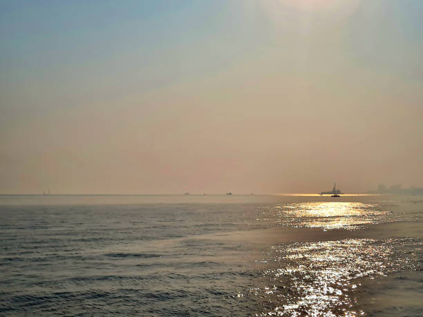 A serene seascape at sunset with a hazy sky and shimmering water reflecting the sun's glow. A distant boat is visible on the horizon, adding to the tranquil atmosphere.