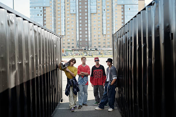 Group of friends standing casually between two black shipping containers with an apartment building in background. They exhibit relaxed and confident postures without looking at camera