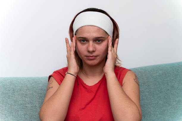 Young woman sitting comfortably on a sofa, wearing a stylish headband, applying face cream to her skin while looking at the camera