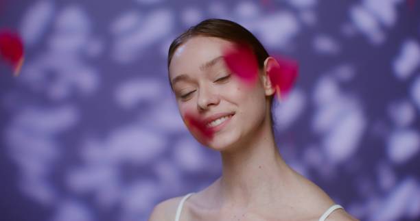 Beautiful young Caucasian model without makeup, with tied hair, smiling broadly looking into the camera while standing against the background of falling rose petals in the studio. 4K video, red komodo