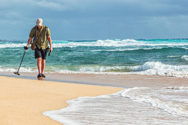 Photo of a man walking along the surf of a remote Hawaiian beach with a hand-held metal detector, searching for lost treasures.