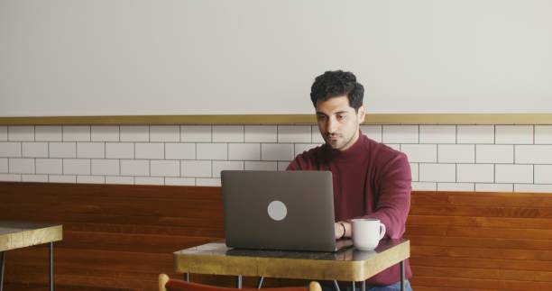 istock Young man uses a laptop sitting at a small table in a cafe, working remotely 2202159149