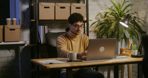 istock Young man works at a laptop sitting at a table in a modern loft-style office 2202159524