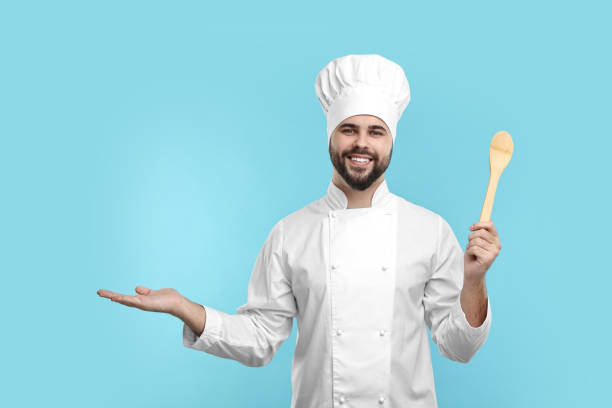 istock Happy young chef in uniform holding wooden spoon on light blue background 2202160824