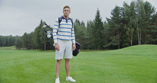 Young blond man in stylish sportswear looking at the camera, and smiling while standing on a well-groomed golf course and holding game equipment in a bag behind his back