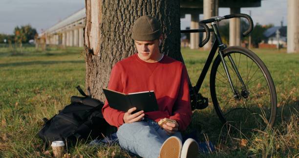 istock A young man reads a book and eats sandwich, sitting on the ground under tree 2202166713