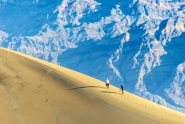 Stovepipe Wells, California, USA – February 14, 2025: Two young adults are hiking in the sand dunes at Mesquite Flat in Death Valley National Park.  The Grapevine Mountains are in the background.