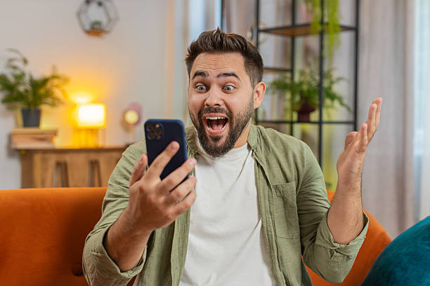 istock Surprised Caucasian brunette man using smartphone winning lottery sitting on sofa couch at home 2202174728