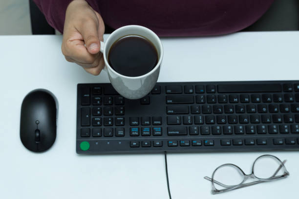 A top-down view of a hand holding a cup of coffee, surrounded by computer accessories and glasses. The setup suggests a workspace or home office environment focusing on comfort and productivity.