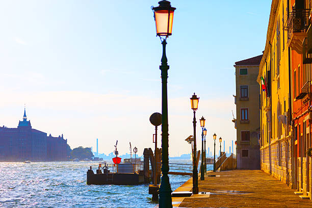 Sunlight bathes a Venetian promenade, where vintage lamp posts line the waterfront beside historic buildings. Tranquil canal reflects the golden hues of sunset, capturing timeless charm of Venice